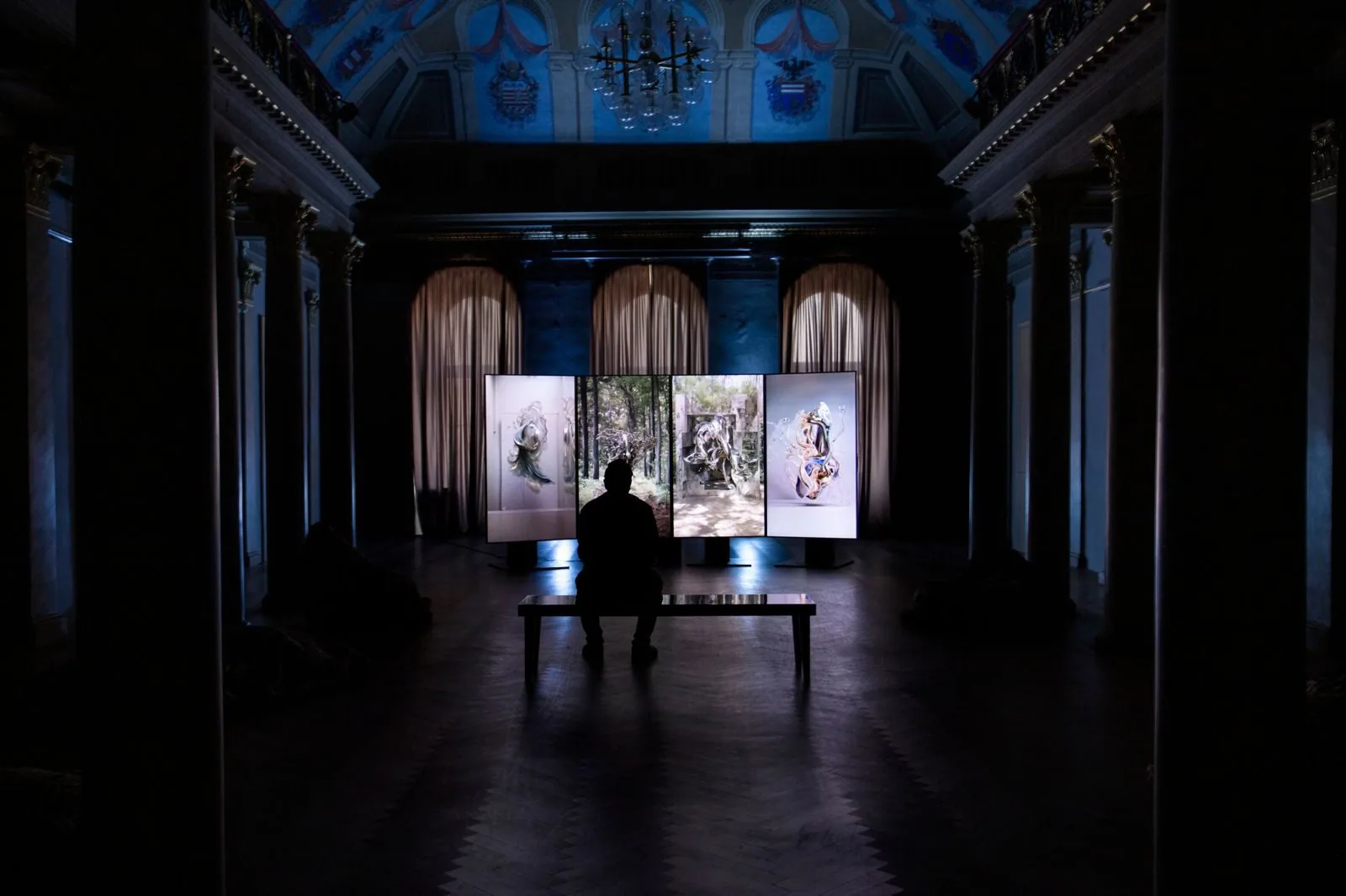 A seated visitor faces a multi-screen installation in a dark historic room